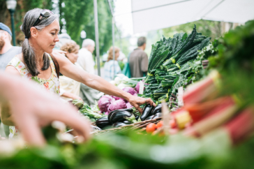 A middle-aged white woman shops at a farmers' market, picking produce from a stall
