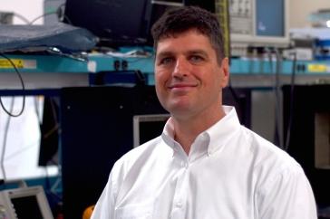 Nathan Schwadron sits in a room full of computer equipment and smiles at the camera.