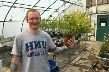 温室里的男学生拿着一株植物. 他的t恤上写着主要研究研究生院.