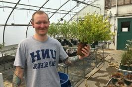 Male student in greenhouse holds a plant. His t-shirt says UNH Graduate School.